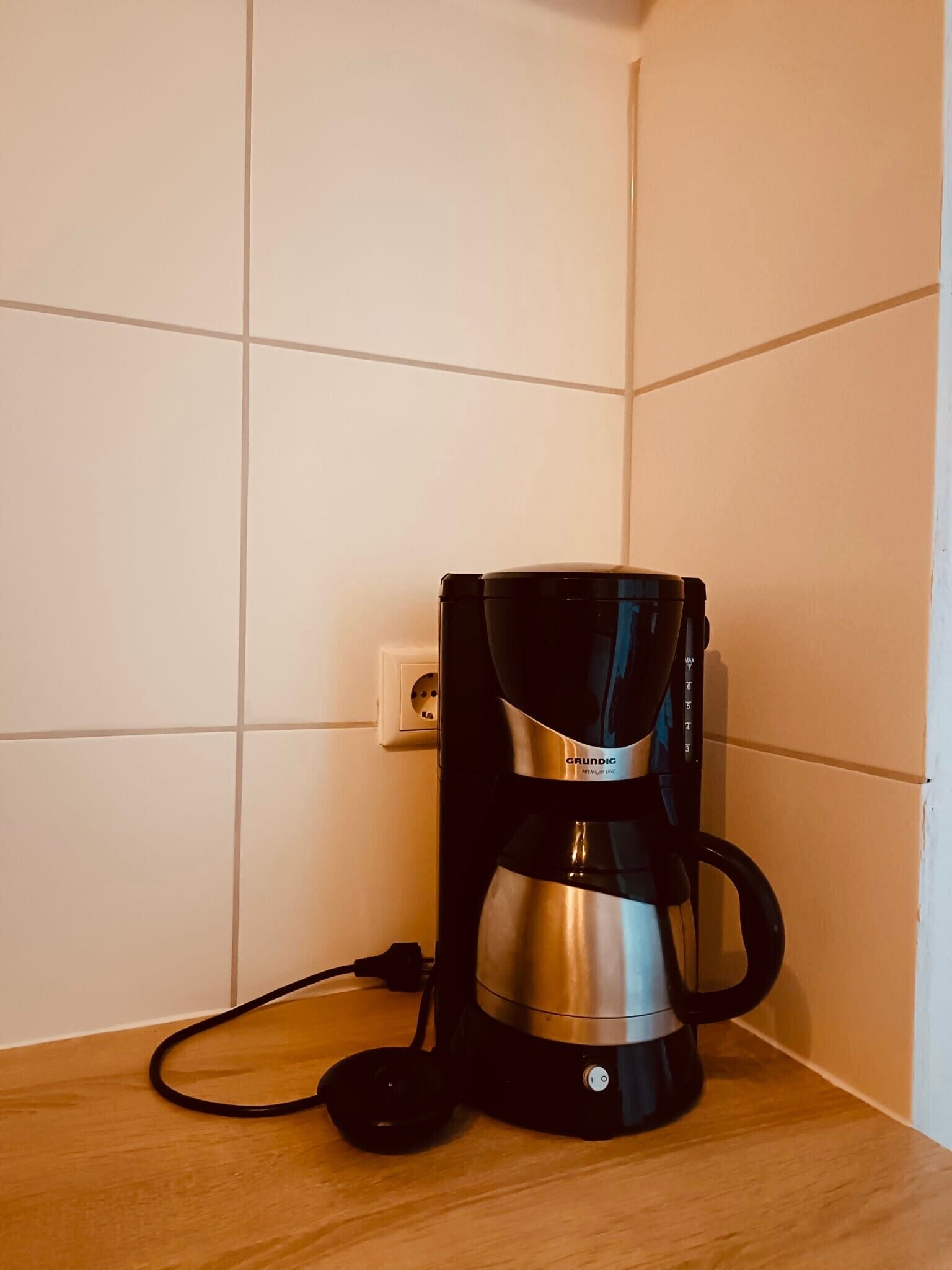 A black coffee maker on a wooden countertop in a corner with white tiled walls.