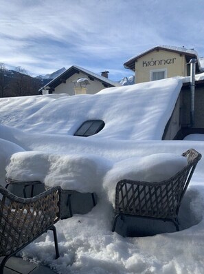 Un espace extérieur enneigé avec deux chaises en osier et, au fond, un bâtiment portant l’inscription « kronner ».