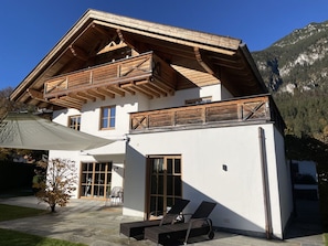 A two-story chalet with a wooden balcony, large windows, and a patio area with lounge chairs. - Apartment 'Ferienwohnung Max 3' with Mountain View, Private Terrace and Wi-Fi (Garmisch-Partenkirchen)