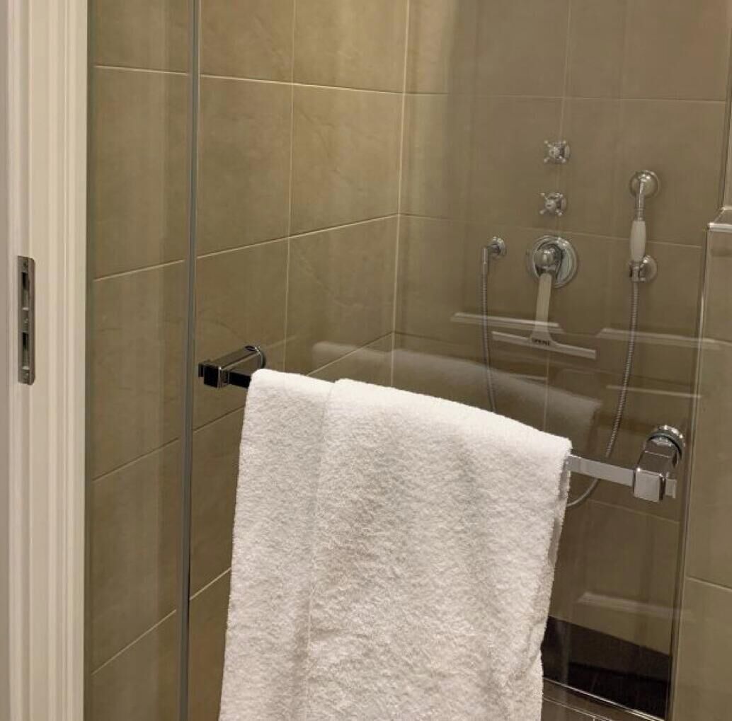Ducha con paredes de vidrio, una toalla blanca en un perchero, un cabezal de ducha manual y un grifo empotrado.