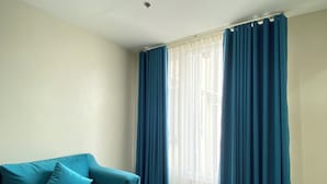 Ein Zimmer mit einem Salbeigrünfarbenen Sofa, einer Deckenleuchte und einem Fenster mit blauen Vorhängen.