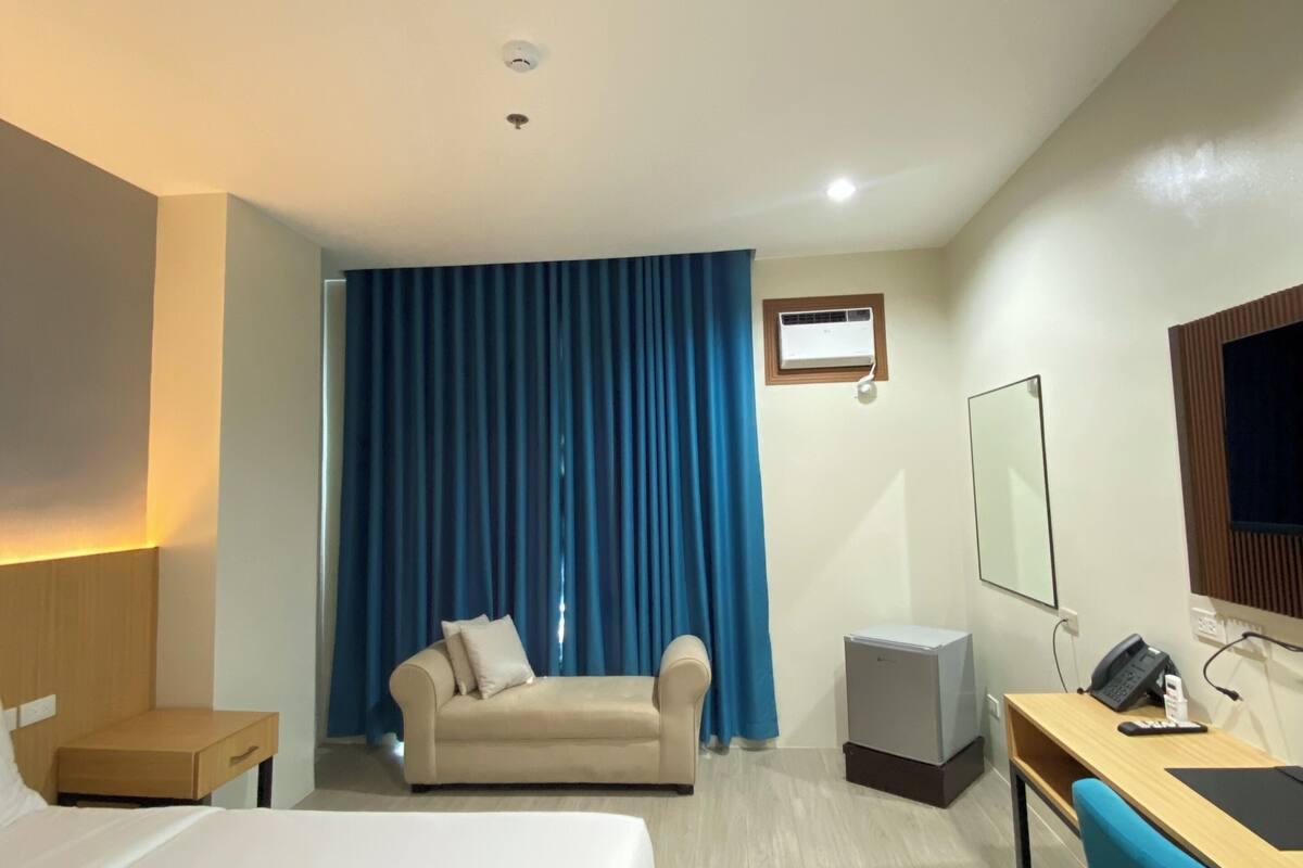 A hotel room with a bed, a desk with a chair, a sofa, a TV, and a window with curtains.