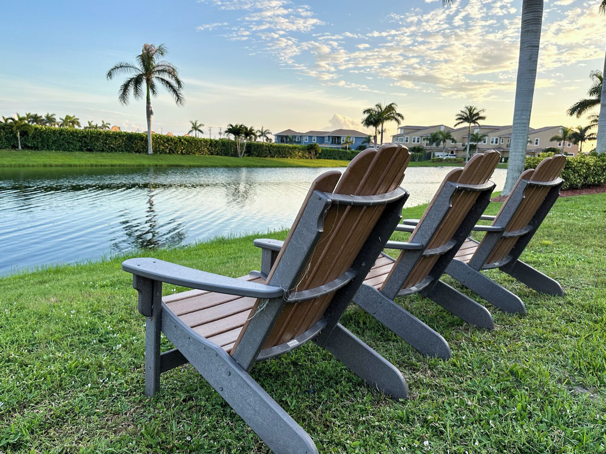 Three Adirondack chairs by a lake with a view of houses and palm trees in the background.