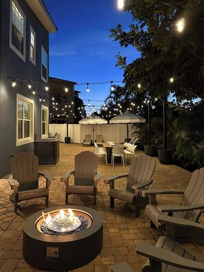 An outdoor patio area with Adirondack chairs, a fire pit, string lights, and umbrellas.