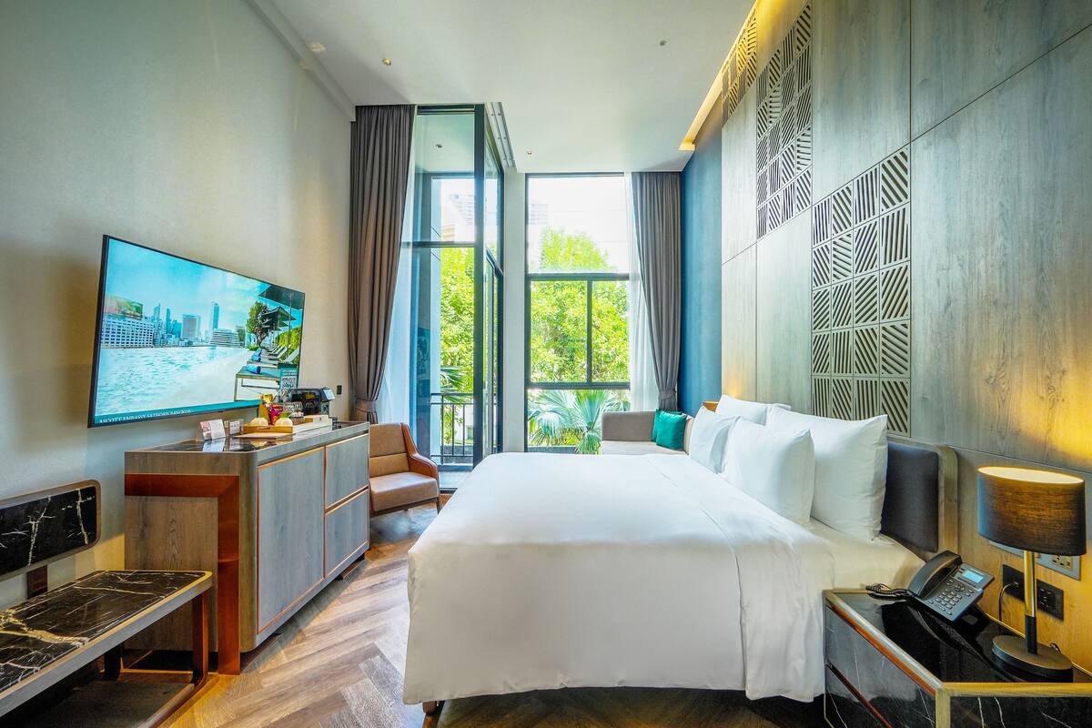Ein modernes Hotelzimmer mit einem großen Bett, einem Schreibtisch mit Stuhl, einem Fernseher und Blick auf Grün durch große Fenster.