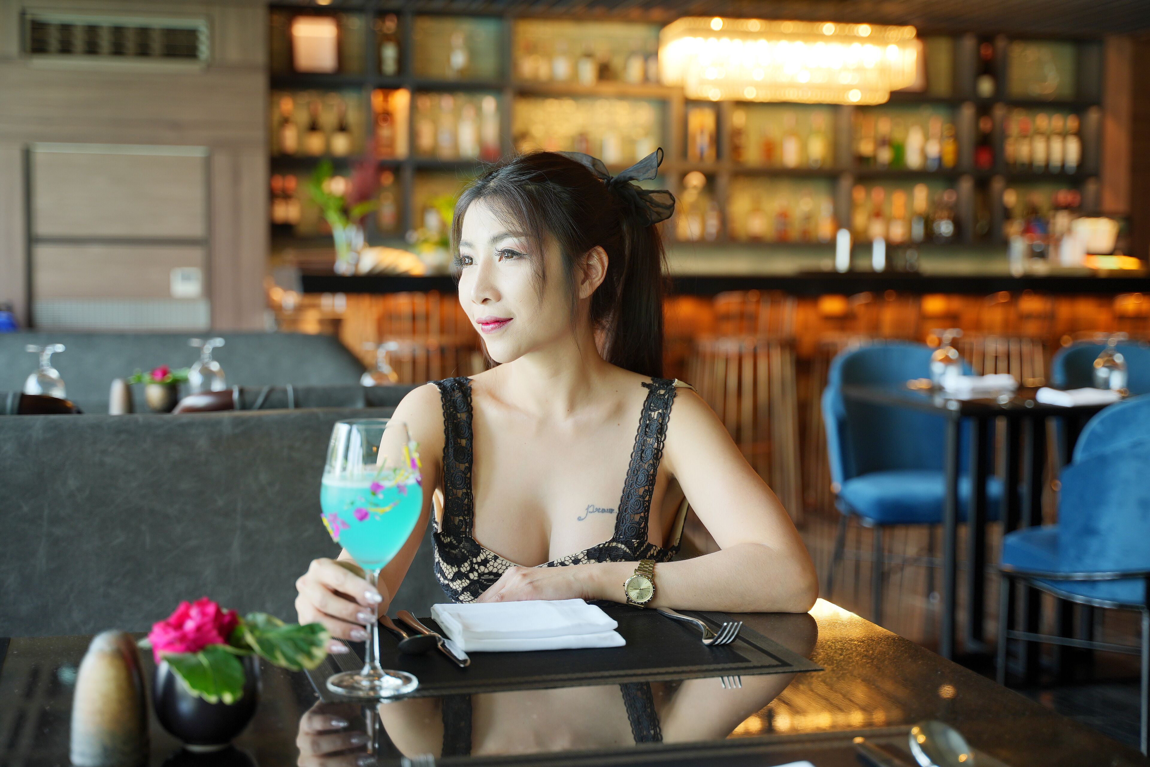 A woman seated at a table in a bar area with a drink, a menu, and a vase with a flower.