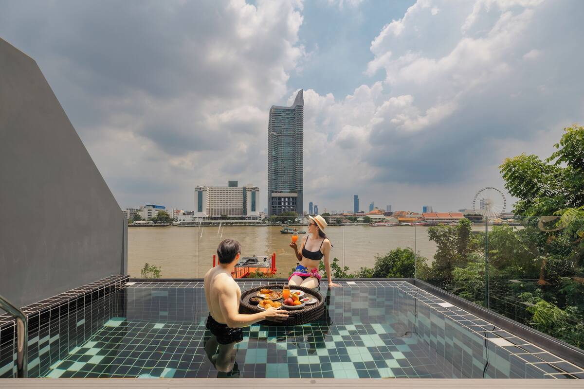 Ein Pool auf dem Dach mit Blick auf die Stadt, ein Esstisch mit Speisen und eine Person, die eine andere bedient.