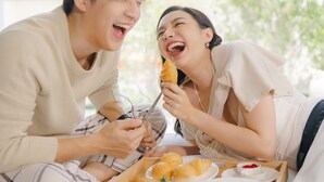 A couple sitting on a bed with a tray of food, including a sandwich and salad, in a room with a city view. - Ten Six Hundred, Chao Phraya, Bangkok by Preference, managed by The Ascott Limited (Bangkok)
