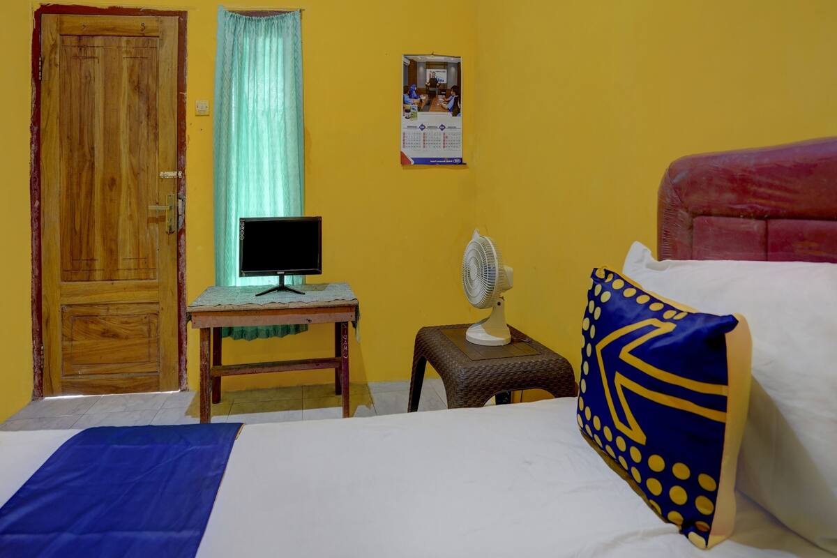 Een kamer met een houten deur, een bureau met een computer, een staande ventilator, een bed met een blauw kussen en een kalender aan de muur.