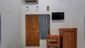 Un dormitorio con un armario de madera, una mesita de madera, una cama, una cortina gris y un televisor montado.