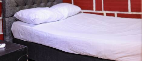 A room with a bed against a brick-patterned wall, a grey upholstered headboard, and a white mattress.