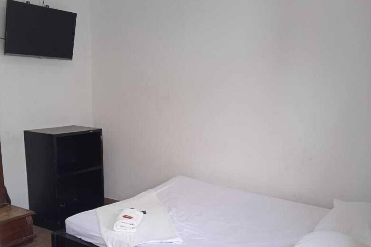Una habitación pequeña y sencilla con una cama, un televisor montado en la pared y un mueble negro pequeño.