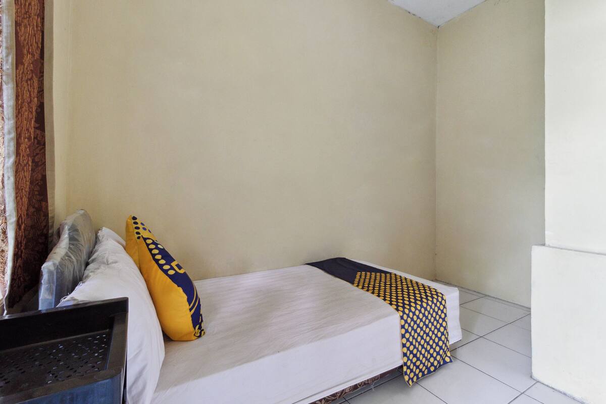 Een kleine eenpersoonskamer met een effen witte muur, een bed met een witte matras en een geborreld deken, een geel kussen, en een zwart object aan de linkerzijde.