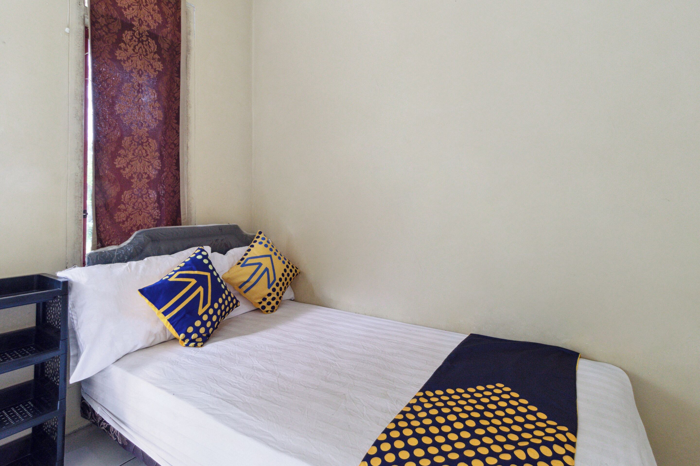 A single bed with white bedding, a black and yellow patterned throw, and two pillows with geometric designs.