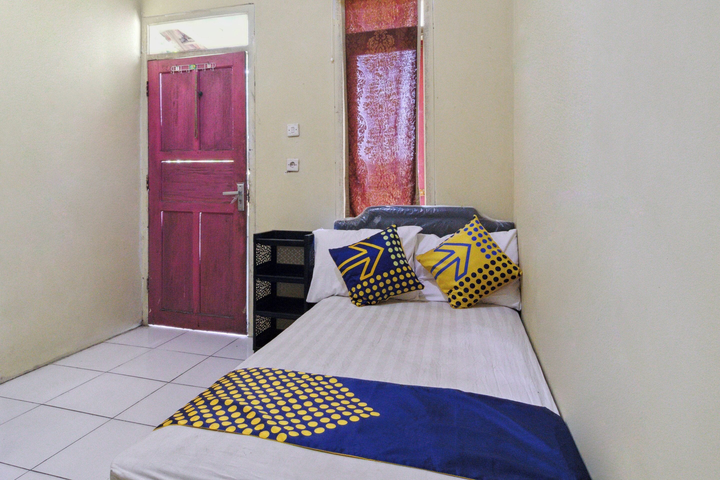 Une petite chambre à coucher, bien rangée, avec un lit simple, une porte rouge et une petite table.
