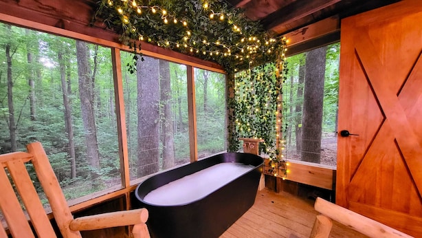 Una cabaña rústica con bañera, sillas de madera y grandes ventanales con vistas a un bosque.