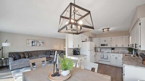 Une cuisine moderne avec des meubles blancs, un coin repas doté d’une table en bois et un salon avec un canapé gris.