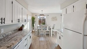 Une cuisine avec des meubles blancs, un plan de travail en marbre, une table à manger avec des chaises et un luminaire suspendu.
