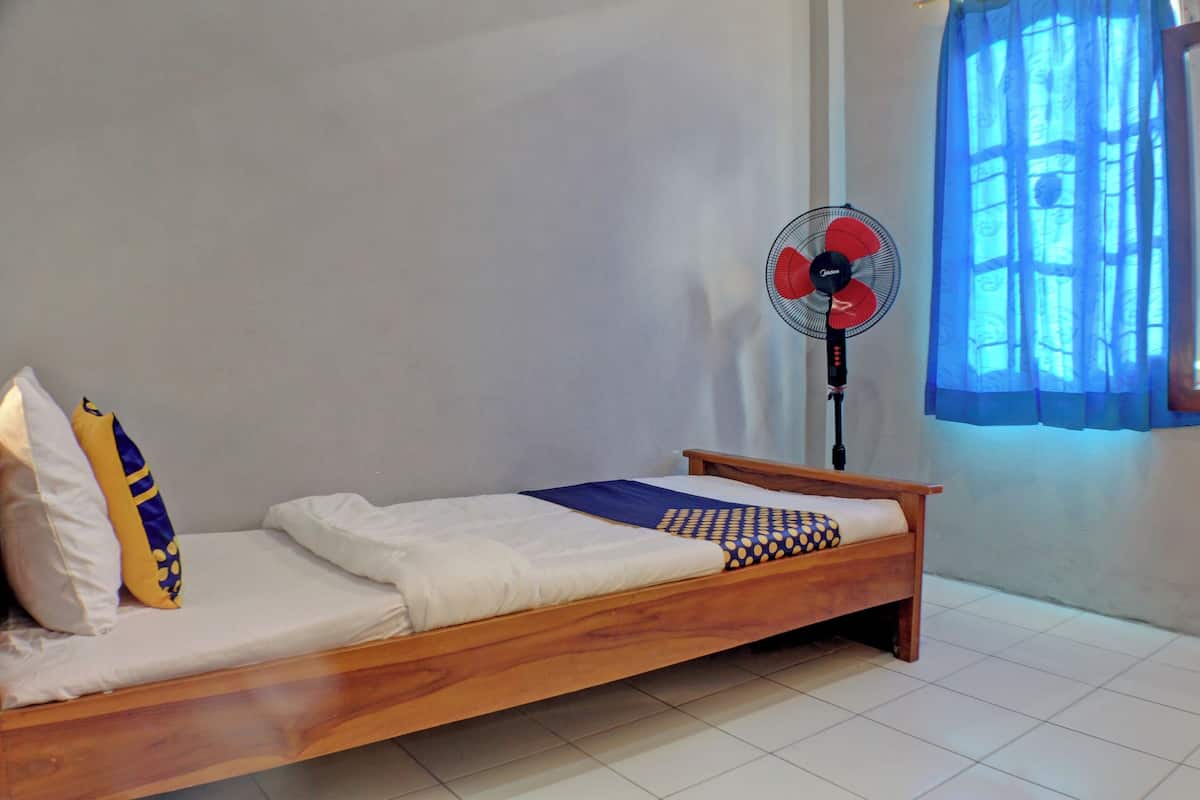 Ein Einzelbett mit Holzrahmen, weißer Bettwäsche und einem blau-gelben Kopfkissen. Ein Standventilator befindet sich rechts neben dem Bett.