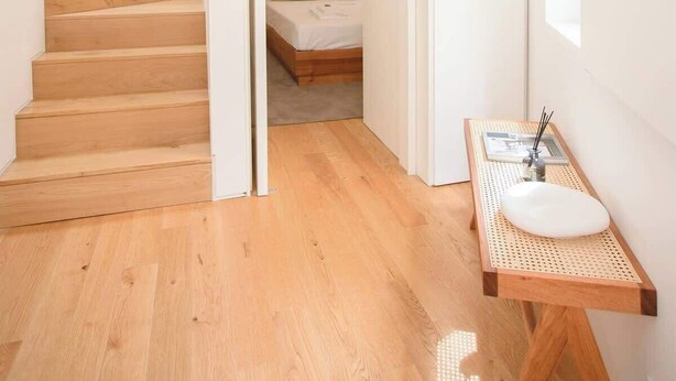 A modern, minimalist bedroom with a wooden bed, a small bedside table with a sink, and a staircase leading to another room.