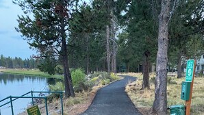 Un sendero pavimentado que lleva a un lago, rodeado de árboles y un cartel que indica reparación de la orilla del río.