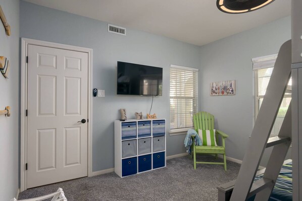 Una habitación infantil con una puerta blanca, un televisor montado en la pared, una silla verde y un mueble de almacenamiento con cajones azules.