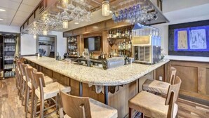 A bar area with a granite countertop, multiple bar stools, hanging wine glasses, and a variety of liquor bottles. - Luxury Condo on Lake Travis with Scenic Views, 3 Pools, Hot Tub, Gym, Tennis, Sauna, Pickleball (Lago Vista)