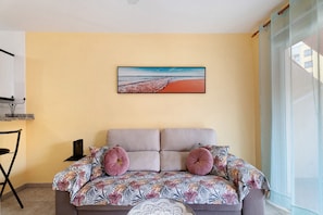Une pièce avec un canapé-lit, un tableau représentant une plage, un fauteuil et une fenêtre donnant sur un paysage agréable.