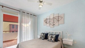 Une chambre à coucher comprenant un lit, une table de chevet, une étagère fixée au mur, un ventilateur de plafond et une vue sur un balcon.