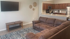 Una sala de estar con un sofá seccional marrón, un pequeño banco de madera, un tapete estampado, un televisor de pantalla plana montado en la pared y una zona de cocina con armarios de madera y electrodomésticos de acero inoxidable.