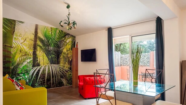Una sala de estar moderna con un sofá rojo, un sillón amarillo y una mesa de comedor de vidrio con sillas. Un televisor grande está montado en la pared, y un mural tropical adorna el espacio.