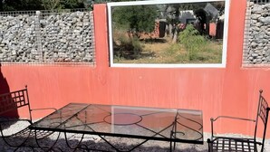 Ein Tisch mit Glasplatte und Metallstühle, auf Kiesboden, vor einer roten Wand mit einem gerahmten Bild.