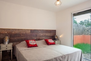 Une chambre à coucher avec une tête de lit en bois, un lit avec deux oreillers rouges, une table de chevet sur laquelle se trouve un casque, et une grande fenêtre donnant sur un paysage verdoyant.