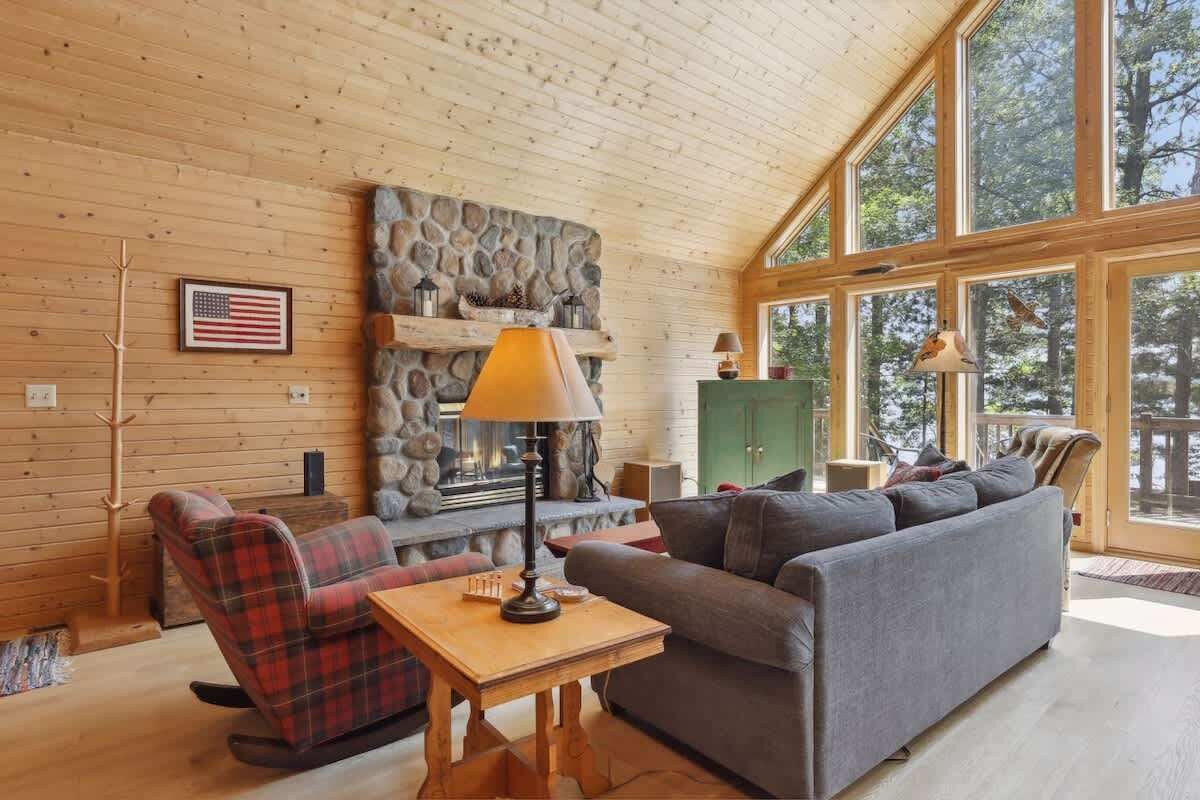 A cozy cabin interior with a stone fireplace, a rocking chair, a sofa, and large windows offering a view of the outdoors.