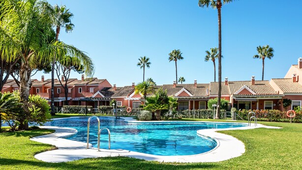 A swimming pool surrounded by palm trees and a row of red-roofed buildings.