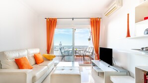 Ein modernes Wohnzimmer mit Sofa, Hocker, Fernseher und einem Balkon mit Stühlen.