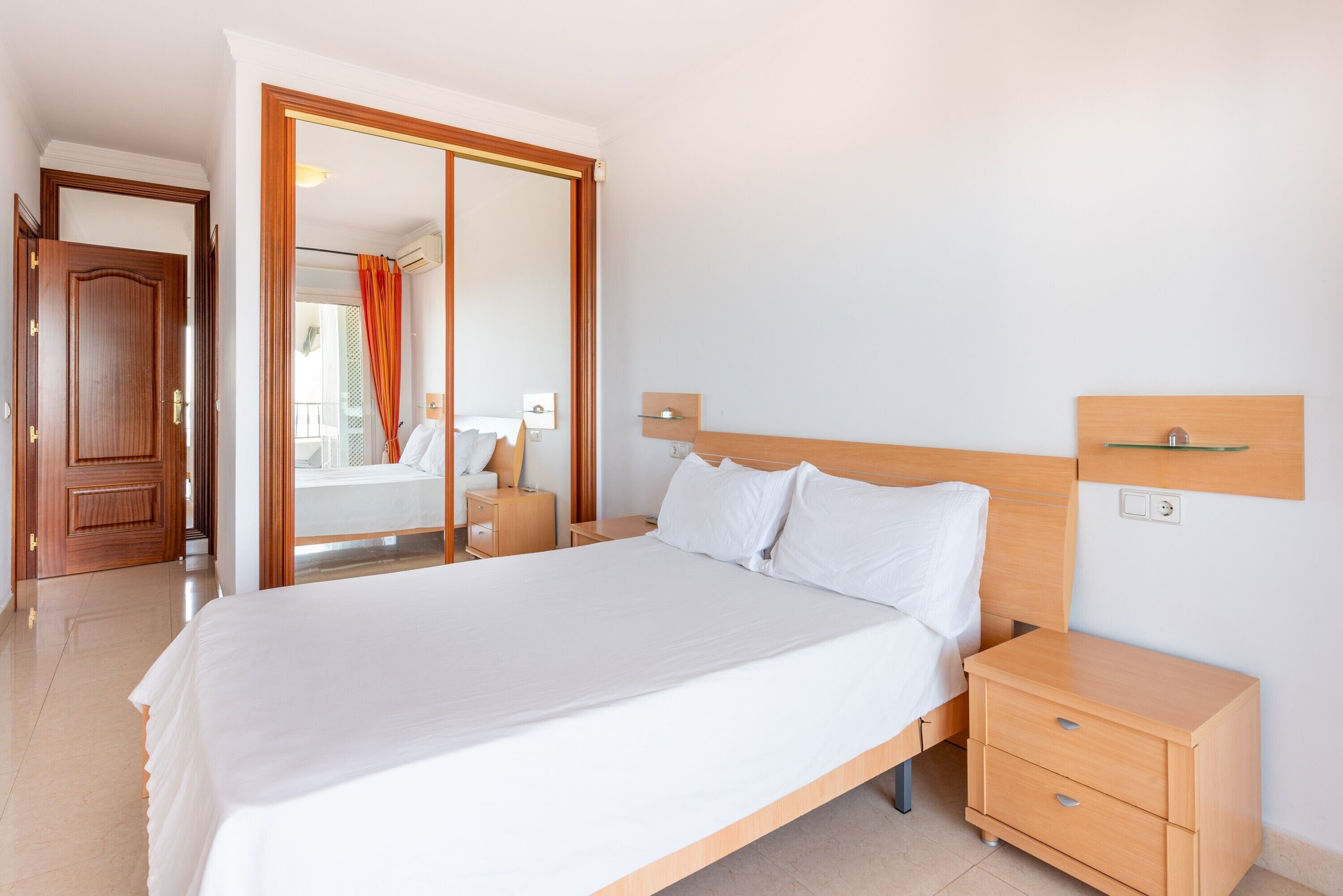 Une chambre d’hôtel avec un grand lit, une tête de lit en bois, des tables de chevet et une vue sur l’extérieur, accessible par une porte coulissante.