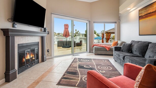 A living room with a fireplace, a television, a sofa, and a large window with a view of a deck and outdoor furniture. - Lakefront Gem • Boat Rental • Sleeps 8 people (Discovery Bay)