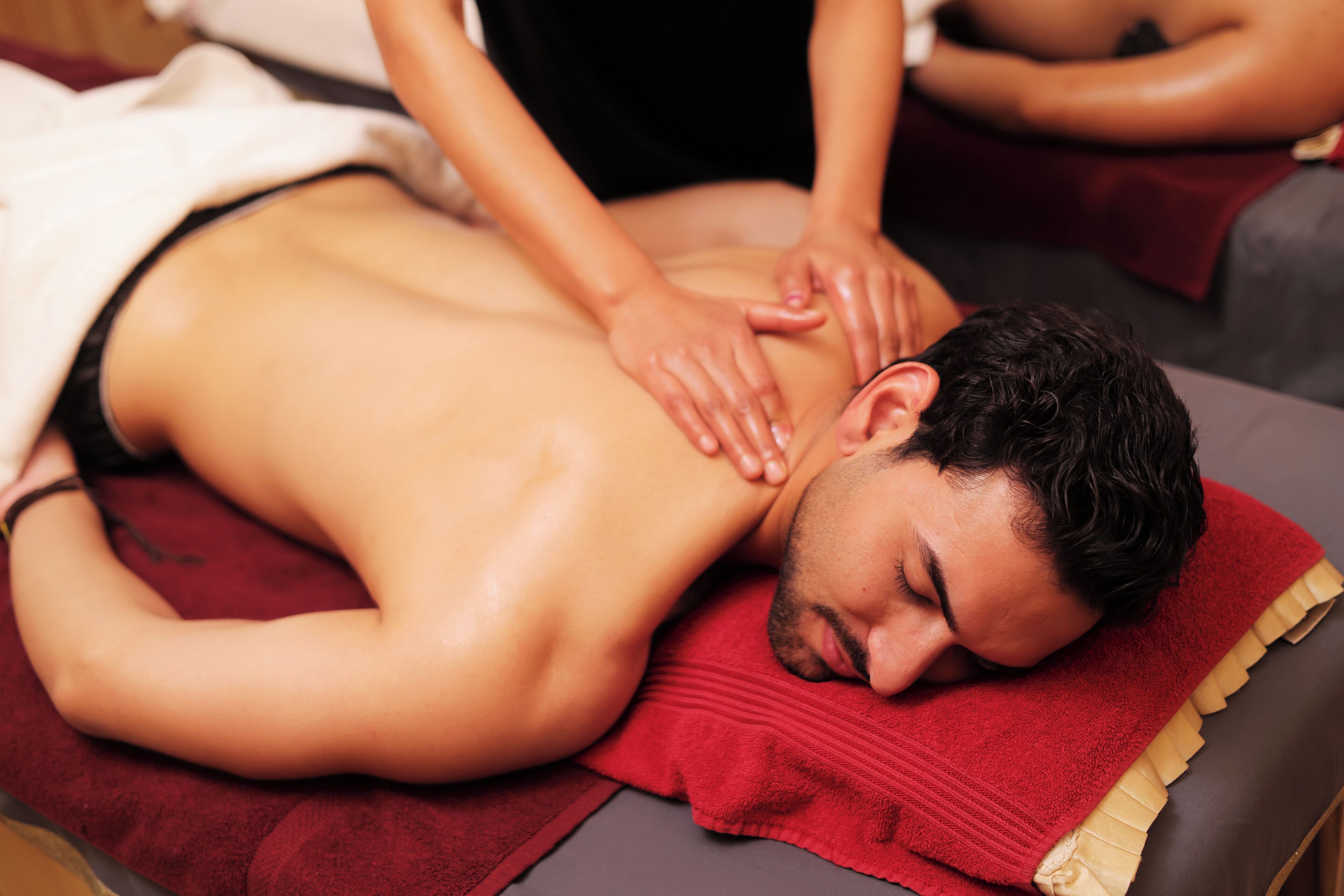 Une personne reçoit un massage du dos sur une table de massage, recouverte d’une serviette rouge et dotée d’un repose-tête en bois.