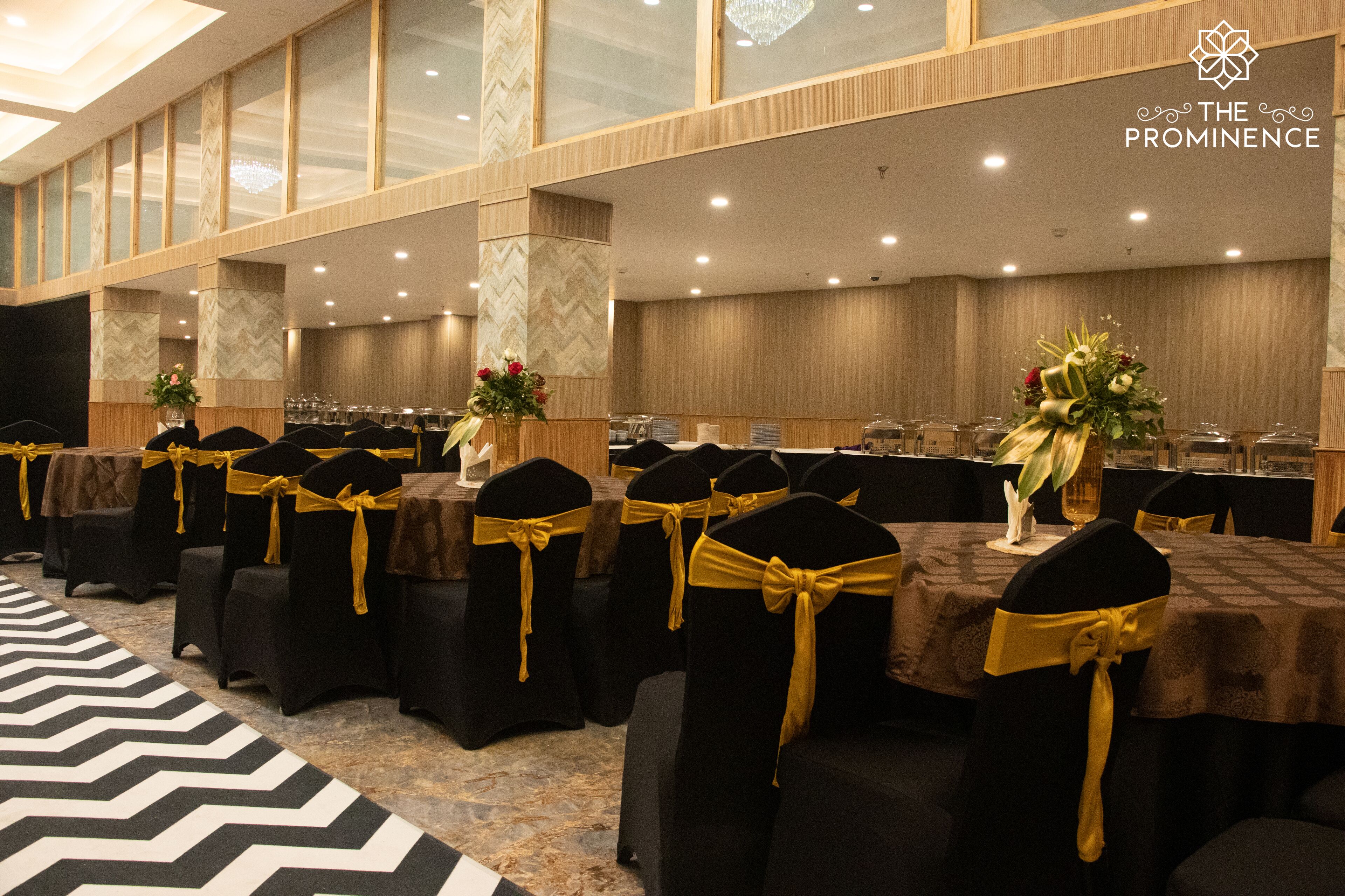 Un salón de banquetes con filas de mesas, cada una con mantel negro y sillas adornadas con cintas amarillas.