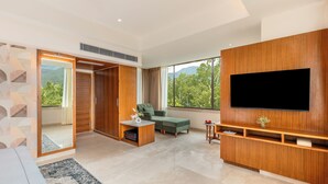 A modern living room with a large TV, wooden furniture, and a view of greenery. - The Prominence Hotel (Dehradun)