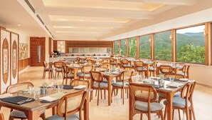 A spacious dining area with multiple tables and chairs, large windows offering a view of the outdoors, and a modern ceiling design. - The Prominence Hotel (Dehradun)
