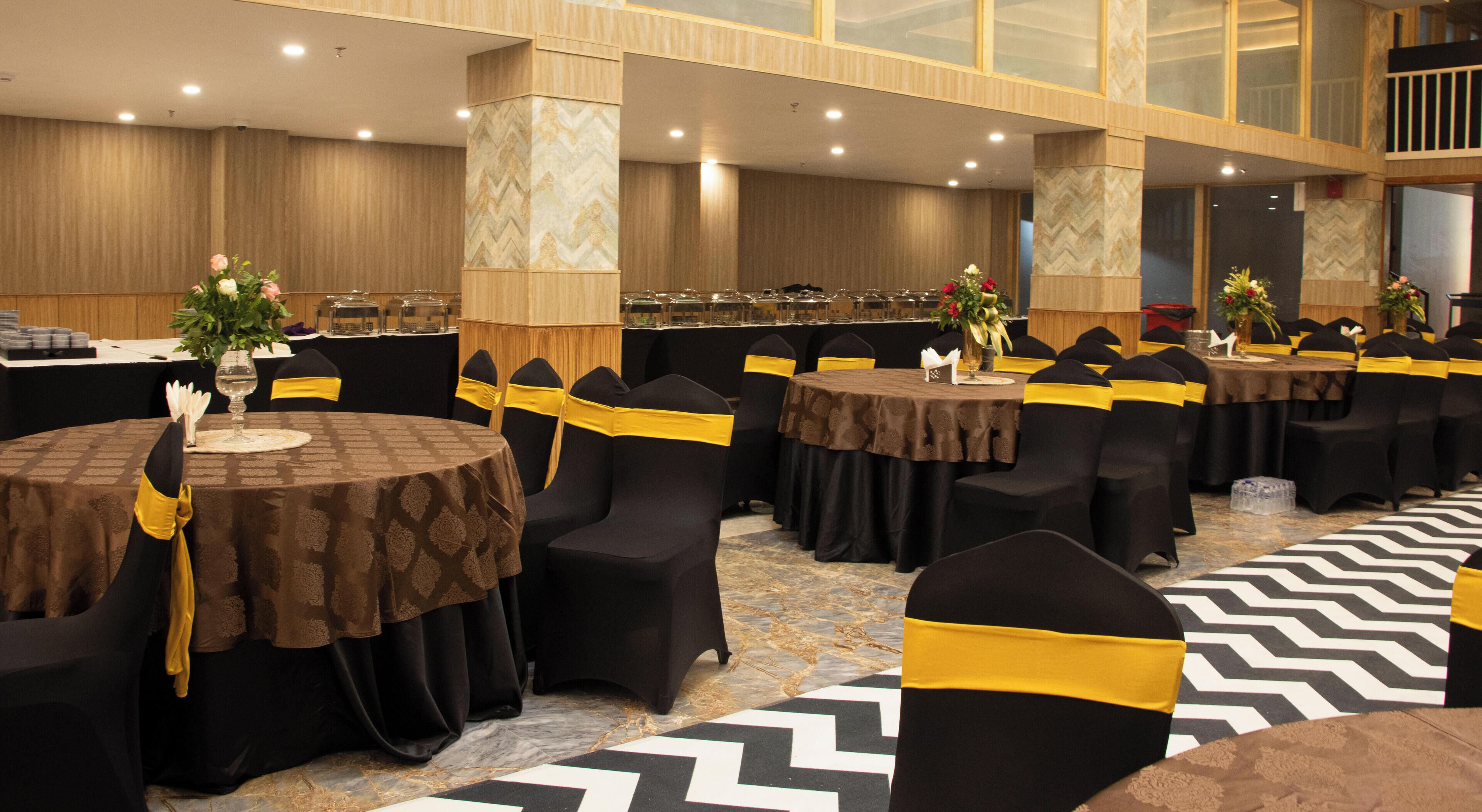 A banquet hall with round tables, black chairs with yellow sashes, and a patterned carpet.