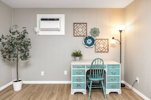 A room with a desk, chair, potted plant, wall-mounted air conditioner, decorative wall art, and a floor lamp.