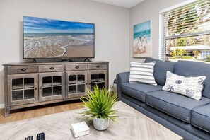 Una sala de estar con televisor, un sofá gris, un mueble y una planta.