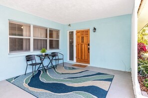 Una habitación con pared azul, puerta de madera, ventana con persianas, una mesita con dos sillas y una alfombra estampada.