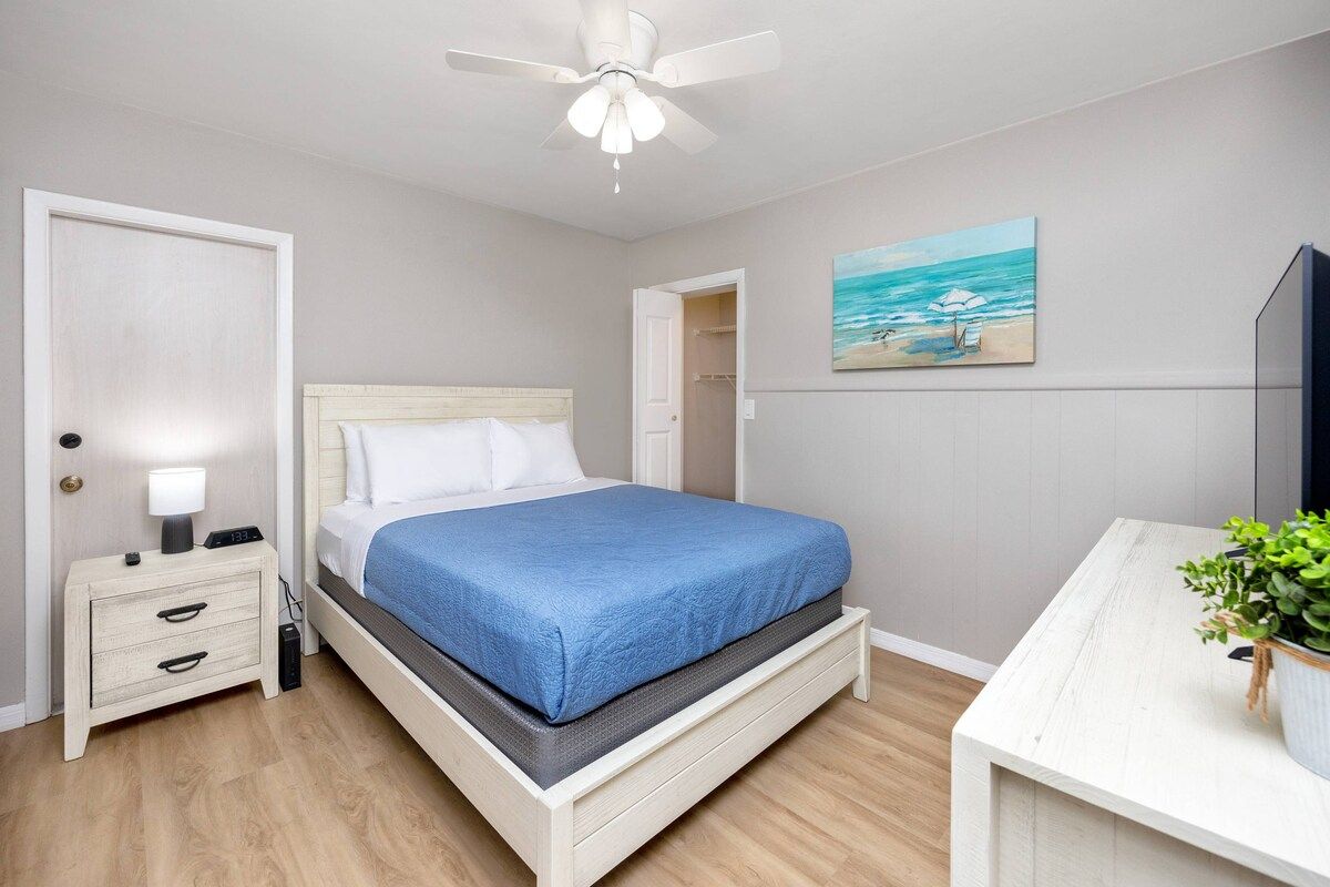 A bedroom with a bed, bedside table, nightstand, ceiling fan, and a painting on the wall.
