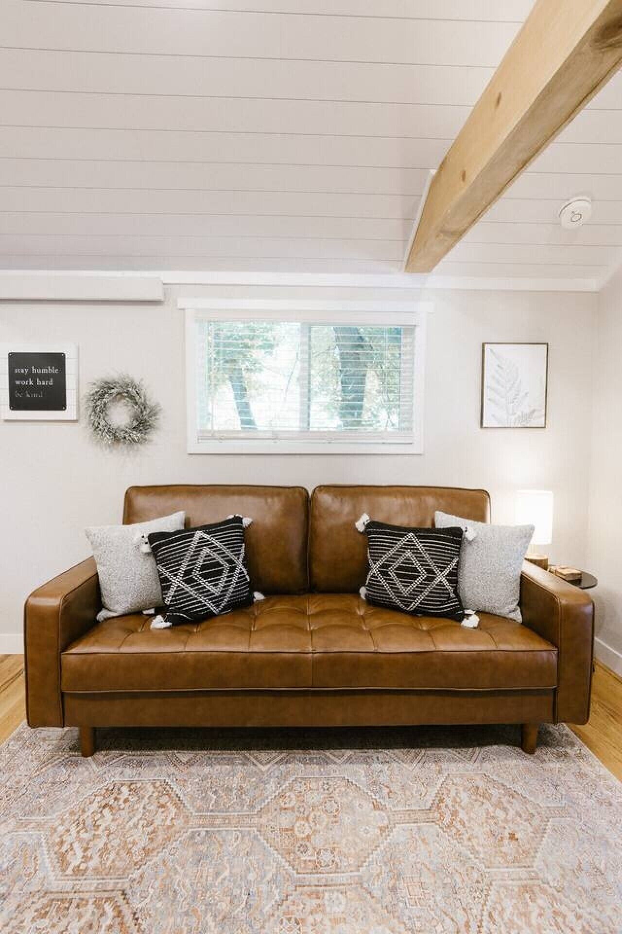 Un canapé en cuir marron agrémenté de coussins à motifs, une couronne décorative et une œuvre d’art encadrée sur un mur blanc.