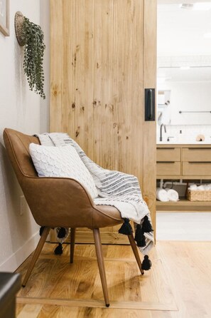 A brown leather chair with a white knitted throw, a wooden door, and a glimpse of a bathroom with a white sink and storage.
