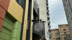 Un edificio de varios pisos con una fachada geométrica distintiva, una entrada visible con un cartel, y vista a la calle con otros edificios y un coche estacionado.